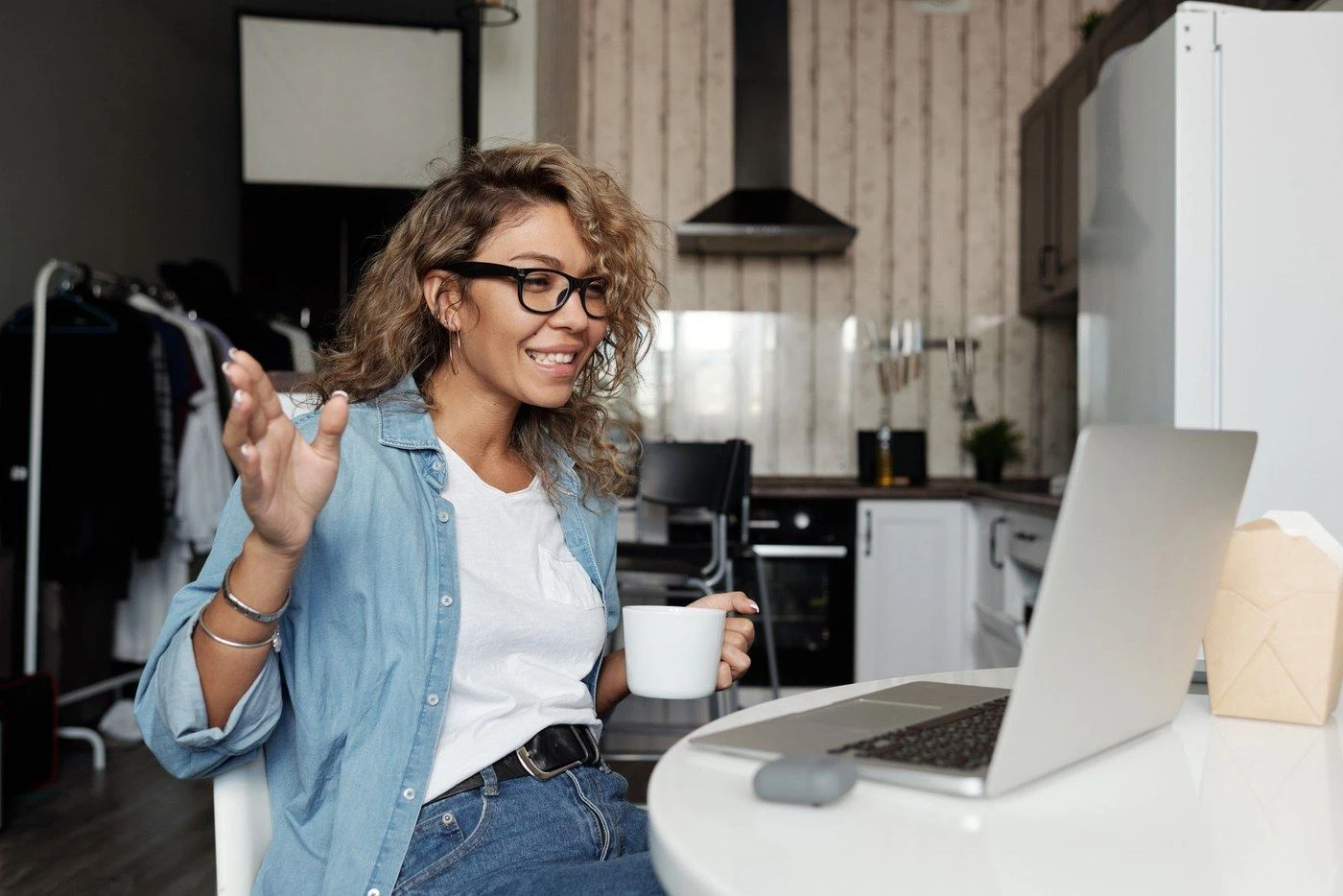 A woman with a coffee talking at her laptop in a kitchen