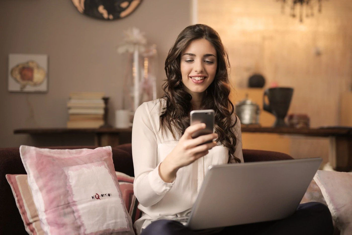 A woman on a sofa smiling at her phone with a laptop on her lap